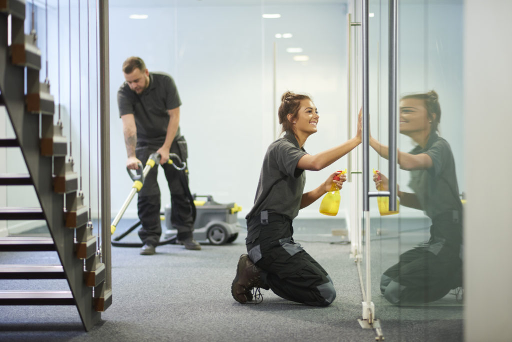 a female cleaning contractor is polishing the glass partition offices whilst In the background a male colleague steam cleans an office carpet in a empty office in between tenants.  .The female is smiling .